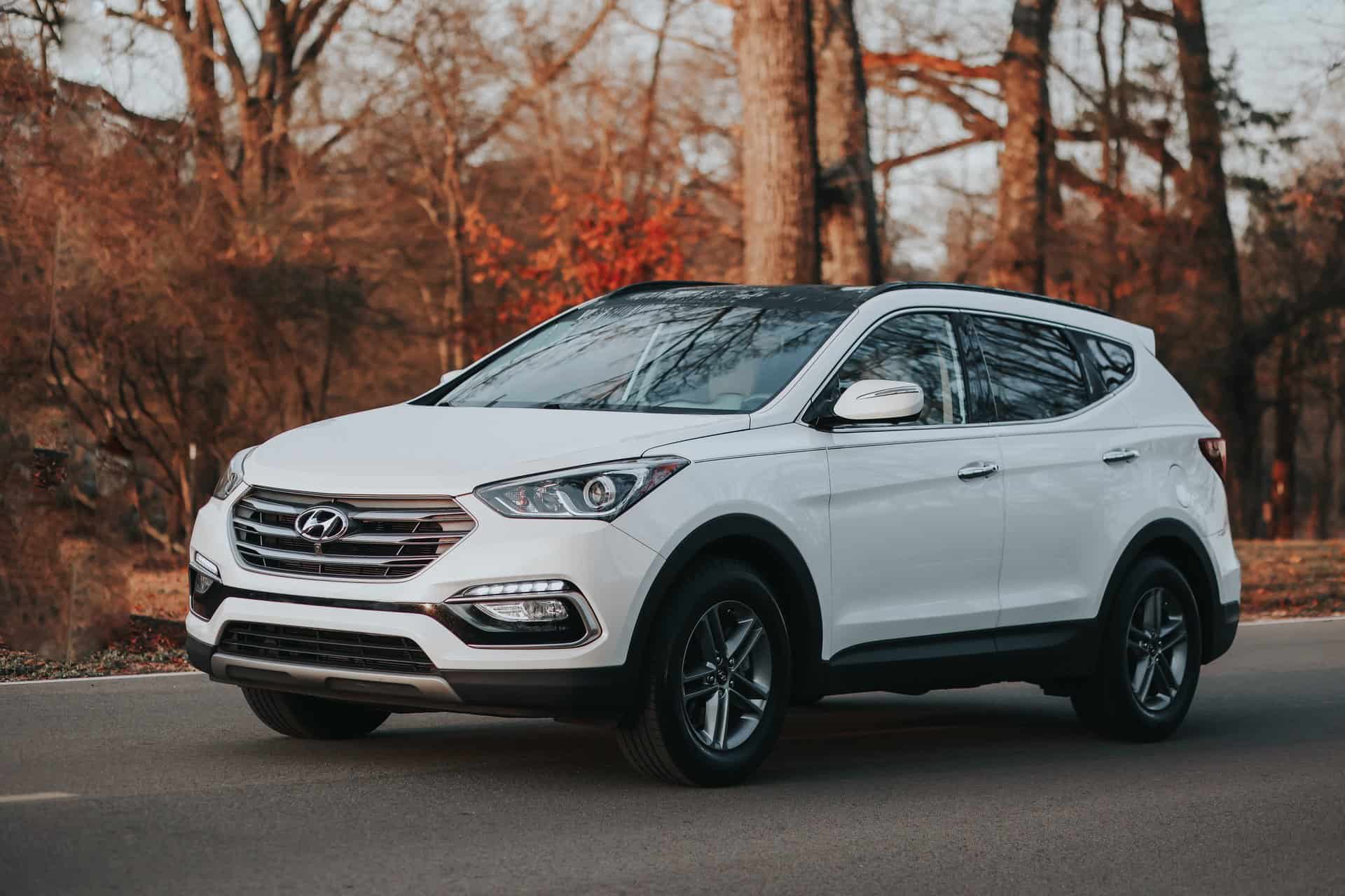 A white hyundai suv parked on the side of an asphalt road surrounded by autumn foliage.