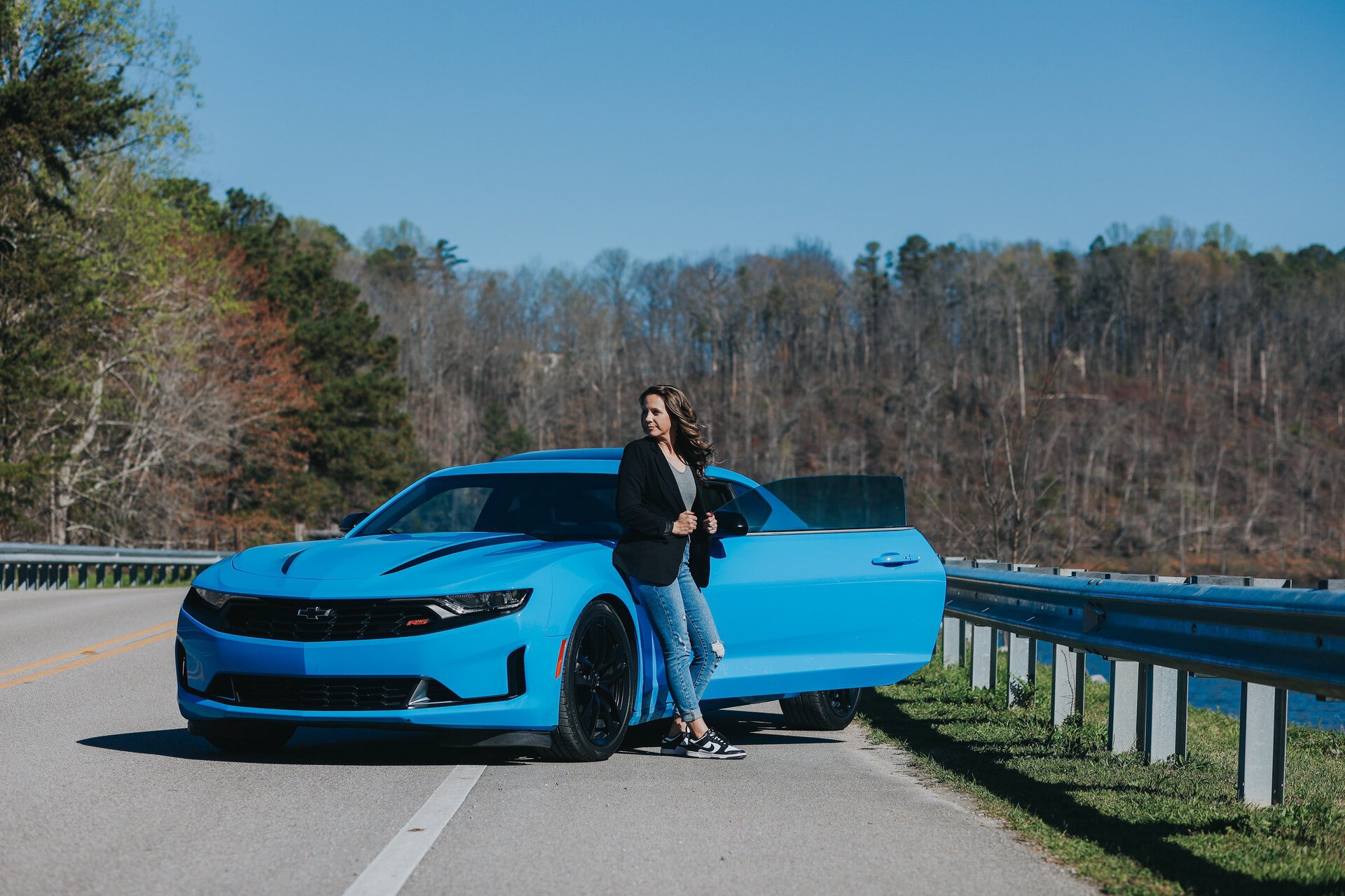 A person stands beside a blue sports car parked on the side of a road with trees in the background.
