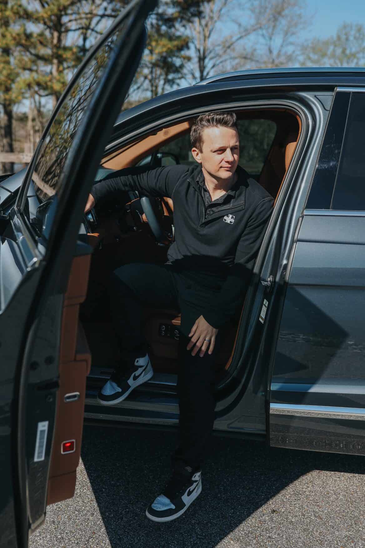 A man in a casual black outfit exits a car, looking to his side with a relaxed posture.