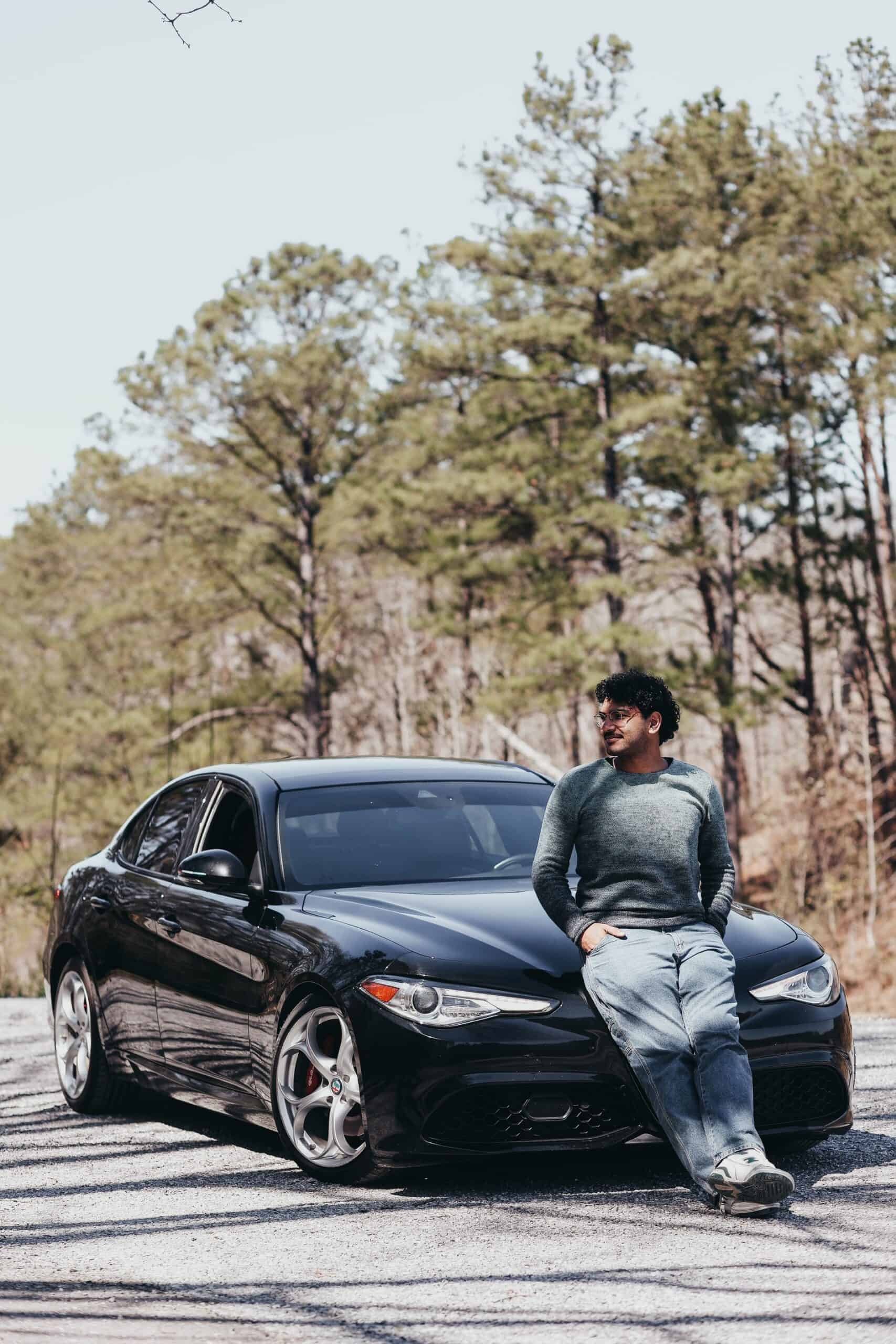 A man wearing sunglasses and casual clothing is sitting on the hood of a black sedan parked on an asphalt road with trees in the background.