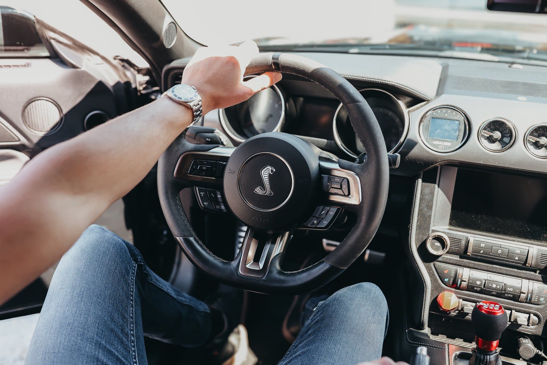 A person holding a steering wheel with their left hand, showcasing a watch on their wrist, inside a car with a manual transmission.