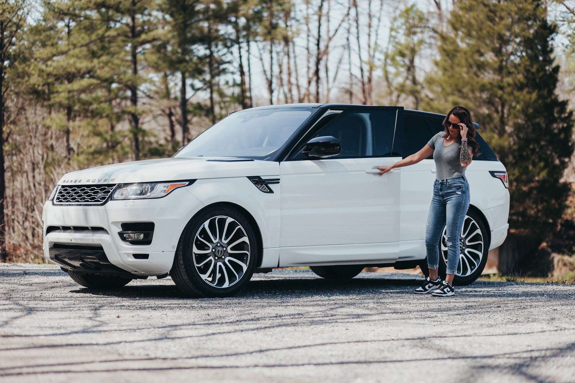 A woman standing by the open door of a white range rover, gesturing with her hand as if in conversation or giving directions.