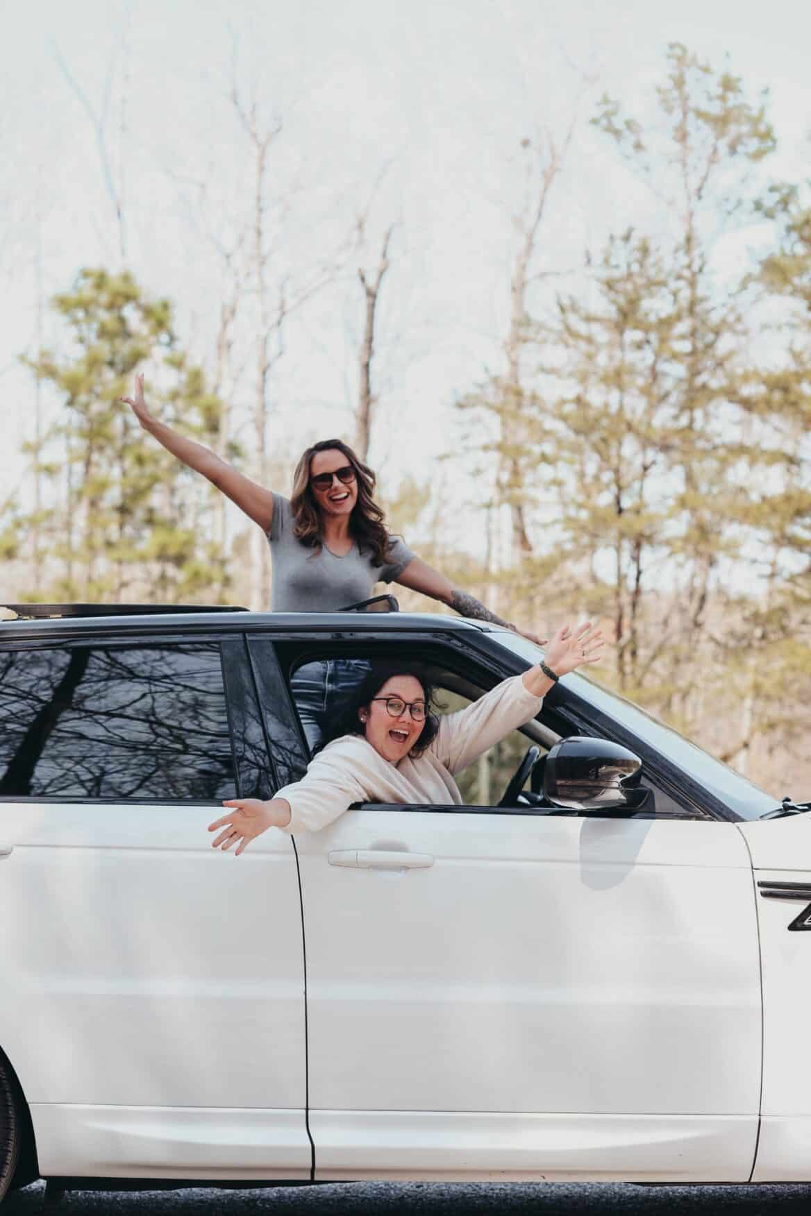 Two individuals are joyfully extending their arms out of the windows of a white car parked in a wooded area.