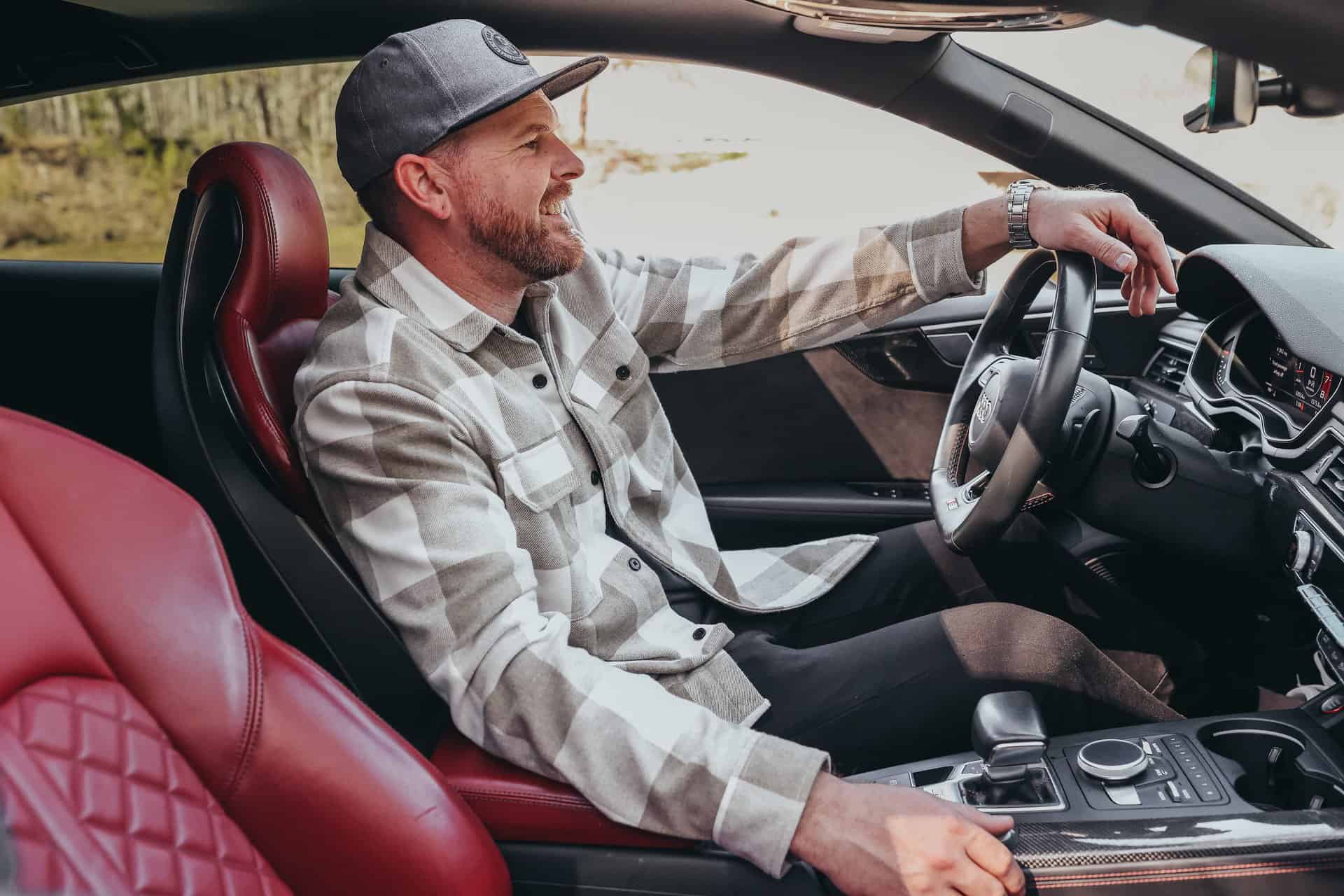 Man in a flat cap and plaid shirt smiling while sitting in the driver's seat of a car with red leather interior.